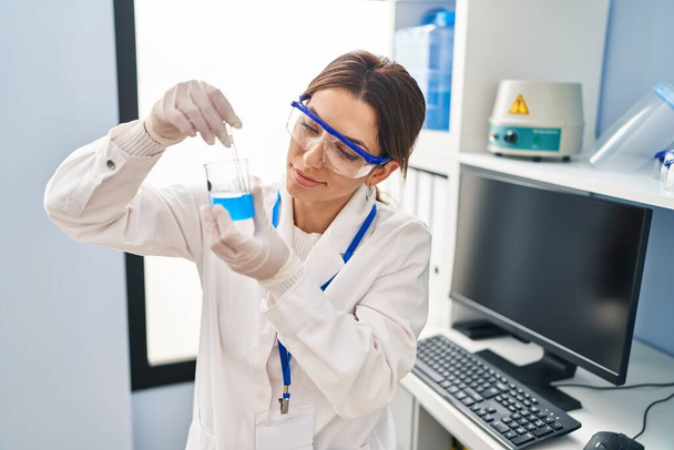 Mujer hispana joven vistiendo uniforme científico mezclando líquido sobre vidrio en laboratorio - Foto, imagen