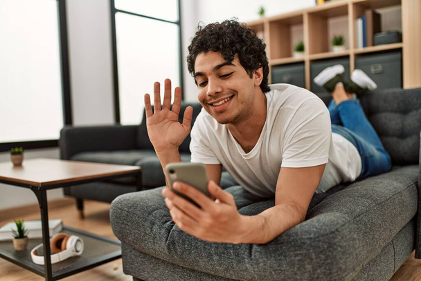 Joven hispano con videollamada usando smartphone tumbado en el sofá de su casa. - Foto, imagen