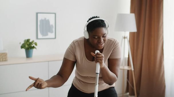 Divertente concetto di lavori domestici. Giovane paffuto africano americano signora indossa cuffie cantare in mop come microfono - Foto, immagini