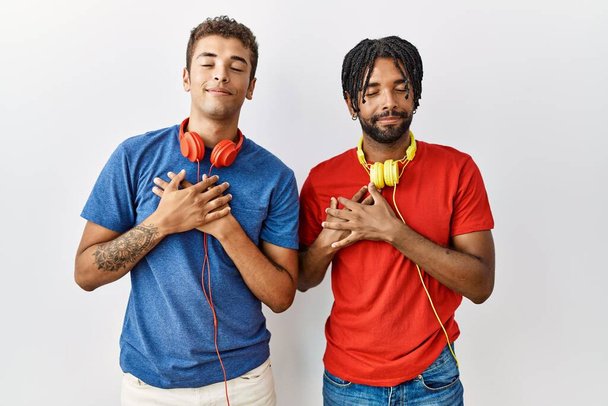 Jonge Spaanse broeders die over een geïsoleerde achtergrond staan en een koptelefoon dragen die glimlacht met handen op de borst met gesloten ogen en dankbaar gebaar op hun gezicht. Gezondheidsconcept.  - Foto, afbeelding
