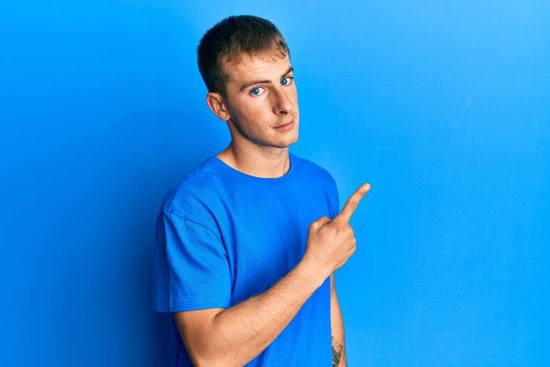 Молодой кавказский мужчина в обычной синей футболке, указывая пальцем в сторону, показывая рекламу, серьезное и спокойное лицо  - Фото, изображение