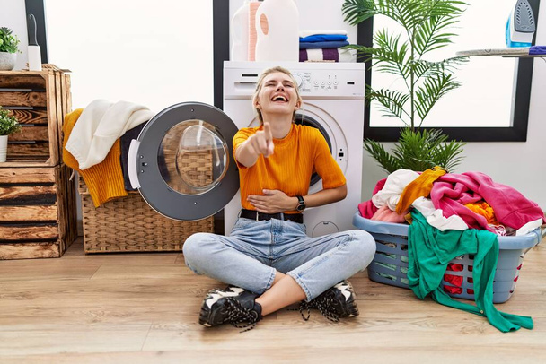 Junge blonde Frau, die Wäsche wäscht, sitzt an der Waschmaschine und lacht dich an, zeigt mit der Hand über den Körper in die Kamera, schämender Ausdruck  - Foto, Bild