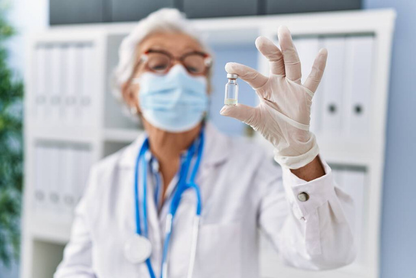Старша сіро-волохата жінка в уніформі лікаря та медичній масці, що тримає вакцину в клініці
 - Фото, зображення
