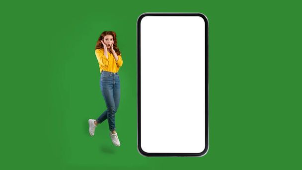 Mujer conmocionada cerca de la pantalla del teléfono móvil en blanco saltando sobre el fondo verde - Foto, imagen