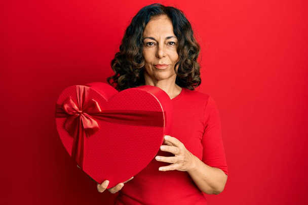 Mujer hispana de mediana edad sosteniendo actitud de pensamiento de regalo de San Valentín y expresión sobria buscando confianza en sí misma  - Foto, imagen