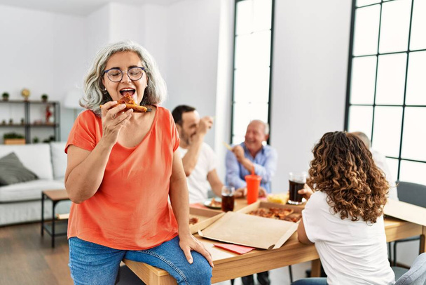 Група людей середнього віку посміхаються щасливо їдять італійську піцу, сидячи на столі вдома
 - Фото, зображення
