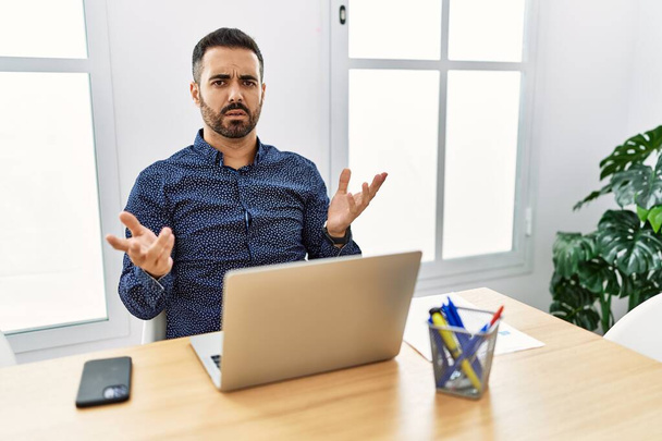 Jovem hispânico com barba trabalhando no escritório com laptop indolente e confuso com os braços abertos, nenhum conceito ideia.  - Foto, Imagem