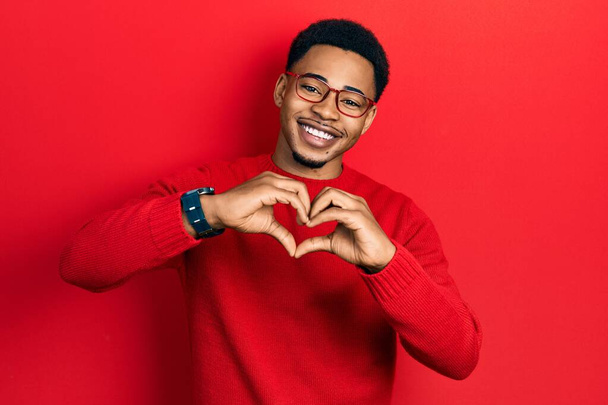Młody, afrykański Amerykanin noszący luźne ubrania i okulary, uśmiechający się w miłości, robiący ręcznie kształt symbolu serca. romantyczna koncepcja.  - Zdjęcie, obraz