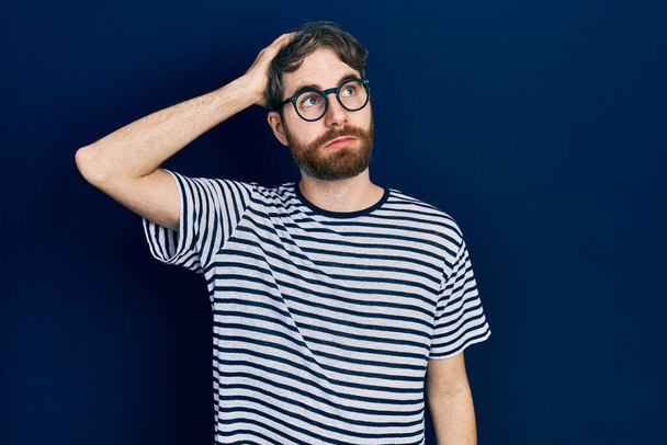 Caucasian man with beard wearing striped t shirt and glasses confuse and wonder about question. uncertain with doubt, thinking with hand on head. pensive concept.  - Photo, Image