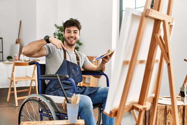 Joven hombre hispano sentado en la pintura en silla de ruedas en el estudio de arte mirando confiado con sonrisa en la cara, señalándose con los dedos orgullosos y felices.  - Foto, Imagen
