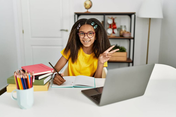 Junge afrikanisch-amerikanische Mädchen, die zu Hause Hausaufgaben machen, lächelnd mit glücklichem Gesicht in die Kamera zwinkern und mit den Fingern ein Siegeszeichen machen. Nummer zwei.  - Foto, Bild