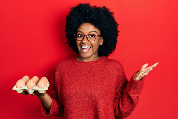 Joven mujer afroamericana mostrando huevos blancos frescos celebrando el logro con sonrisa feliz y expresión ganadora con la mano levantada  - Foto, imagen