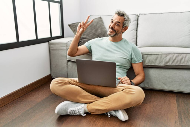 Middle age hispanic man using laptop sitting on the floor at the living room smiling looking to the camera showing fingers doing victory sign. number two.  - Foto, immagini