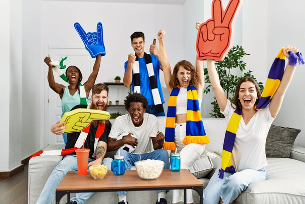 Ομάδα νεαρών φίλων που παρακολουθούν και υποστηρίζουν ποδοσφαιρικό αγώνα στο σπίτι. - Φωτογραφία, εικόνα