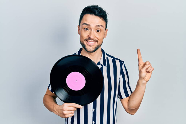 Young hispanic man holding vinyl disc smiling with an idea or question pointing finger with happy face, number one  - Photo, image