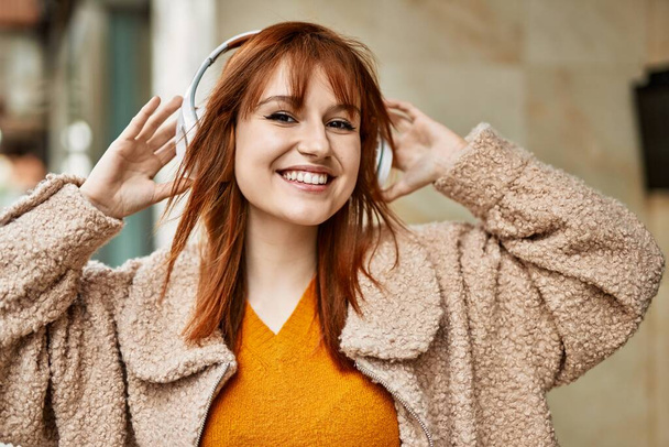 Nuori punapää tyttö hymyilee onnellinen käyttäen kuulokkeet kaupungin. - Valokuva, kuva