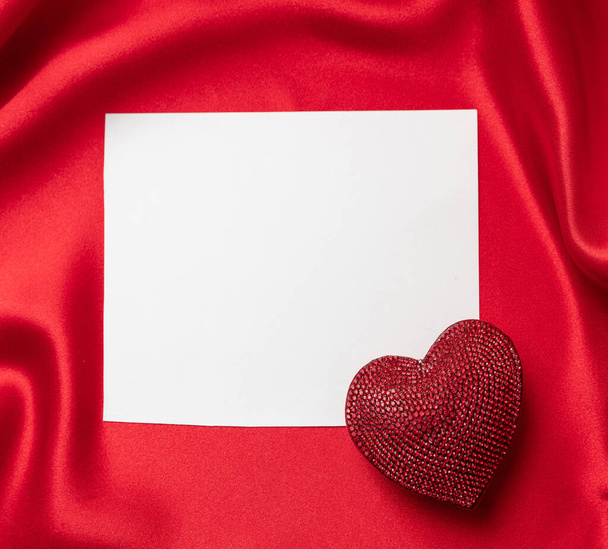 San Valentín con símbolo de amor un color rojo rubí joya en forma de corazón en blanco tarjeta vacía sobre fondo ondulado de seda roja, vista aérea, espacio de copia. - Foto, Imagen