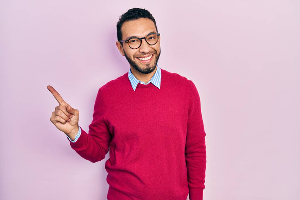 Homme hispanique avec une barbe portant une chemise d'affaires et des lunettes avec un grand sourire sur le visage, pointant du doigt vers le côté en regardant la caméra.  - Photo, image