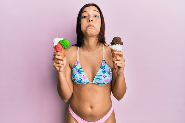 Jeune femme latine portant un bikini tenant une crème glacée gonflant les joues avec un visage drôle. bouche gonflée d'air, attrapant l'air.  - Photo, image