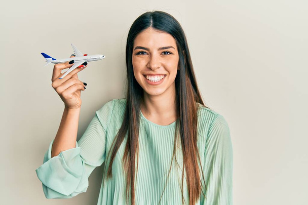 Joven mujer hispana sosteniendo juguete de avión con aspecto positivo y feliz de pie y sonriendo con una sonrisa confiada mostrando los dientes  - Foto, Imagen