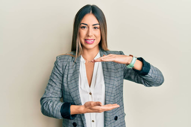 Young hispanic woman wearing business clothes gesturing with hands showing big and large size sign, measure symbol. smiling looking at the camera. measuring concept.  - 写真・画像