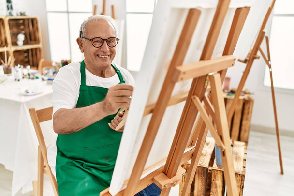 Senior grijsharige kunstenaar glimlachend gelukkig schilderij in kunststudio. - Foto, afbeelding