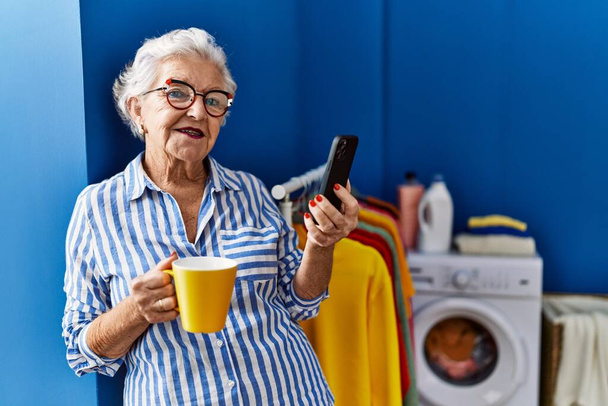 Çamaşır odasında kahve içen, akıllı telefon kullanan yaşlı, kır saçlı kadın. - Fotoğraf, Görsel