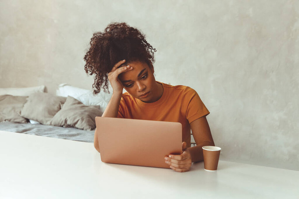 疲れてフラストレーションを感じるアフリカ系の少女がノートパソコンの前の机の上に腰を下ろしていた。フリーランスの感情的な側面 - 写真・画像