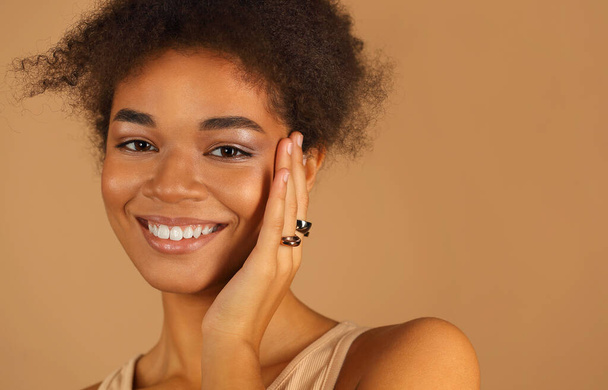 Elegante retrato de perfil de joven sonriente hermosa mujer afroamericana con maquillaje natural demostrando anillos en sus dedos manteniendo las manos en la cara aisladas sobre la pared del estudio beige. Concepto joyería - Foto, imagen