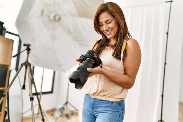 Νεαρή Ισπανίδα φωτογράφος χαμογελά ευτυχισμένη χρησιμοποιώντας κάμερα στο φωτογραφείο. - Φωτογραφία, εικόνα