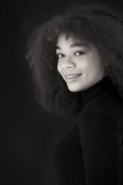 Portrait of attractive smiling curly african-american woman in black clothes looking at camera, posing isolated on dark studio background. Young naturally beautiful african female model without makeup. Black and white - Photo, Image
