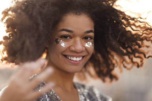 close-up portret van overblijd charmant Afrikaans amerikaanse vrouw met krullend haar en glitter op jukbeenderen glimlachen gelukkig op camera terwijl bewegend dansen buiten, feestelijke sfeer in de lucht - Foto, afbeelding