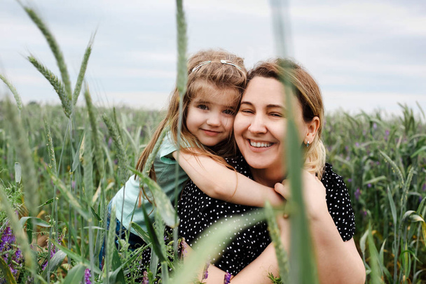 Glückliche Mutter und Tochter mit einem Blumenstrauß, der sich auf der grünen Wiese umarmt. Mutter umarmt kleine Tochter draußen auf einem Feld. Liebe und Familienkonzept - Foto, Bild