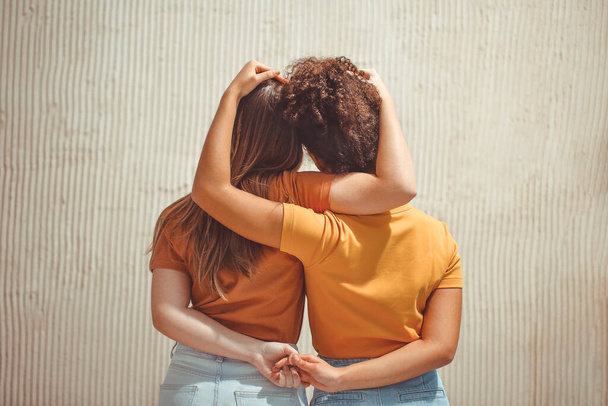 Güçlü bir kadın arkadaşlığı. Arka planda iki genç kız el ele tutuşup dışarıda bej duvarın önünde dikilirken birbirlerine sarılıyorlar. - Fotoğraf, Görsel