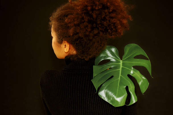Φυσική ομορφιά. Σκούρο πορτρέτο στούντιο της νεαρής αφροαμερικανής γυναίκας με πράσινα φύλλα monstera και κοιτάζοντας κάμερα, σκεπτική αφρικανική θηλυκό εκμετάλλευση τροπικό φυτό απομονωμένο πάνω από το μαύρο τοίχο - Φωτογραφία, εικόνα