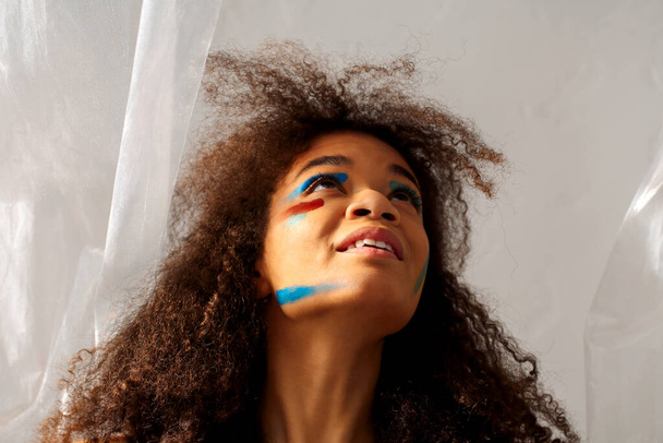 Beautiful happy african american ethnicity woman with creative art makeup looking aside with pleasant smile, cheerful black curly girl with color smears on face posing against gray studio background - Foto, Imagen