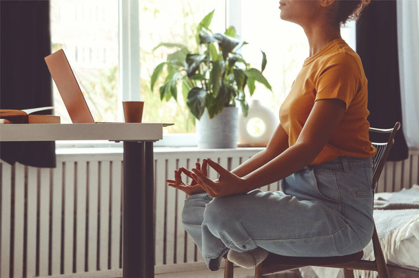 Serbest çalışma sırasında stres atlama. Afro-Amerikan kadın serbest piyango pozunda otururken ve elleri Mudra jesti ile masada açık dizüstü bilgisayarın önünde meditasyon yaparken çekilmiş bir fotoğraf. - Fotoğraf, Görsel