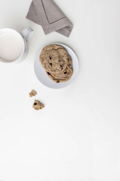 A white mug with milk on a white countertop. There is a napkin next to it and one cookie on a plate. Top view - Photo, image