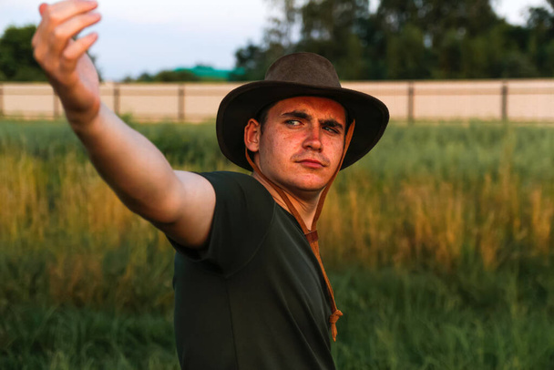 Молодой человек в ковбойской шляпе показывает руки на зеленой траве. Мужчина на голубом фоне неба, на лугу. Концепция сельского хозяйства Вау и весело. Крупный план. Крылья героя, самолет - Фото, изображение