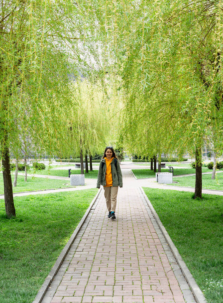 Hymyilevä nuori nainen turisti reppu kävely pitkin kujaa itkien paju nuori vihreä kevät lehtineen, kausi - Valokuva, kuva