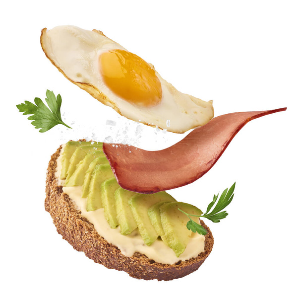 Fresh delicious tasty homemade sandwich with avocado, roasted egg and bacon falling in the air isolated on white background - Photo, Image