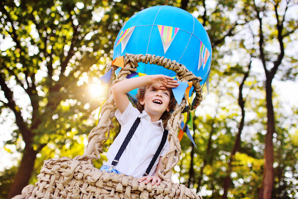 Junge mit lockigem Haar im Korb eines blauen Ballons lächelt und blickt in die Ferne vor dem Hintergrund von Grün und Sonne. - Foto, Bild