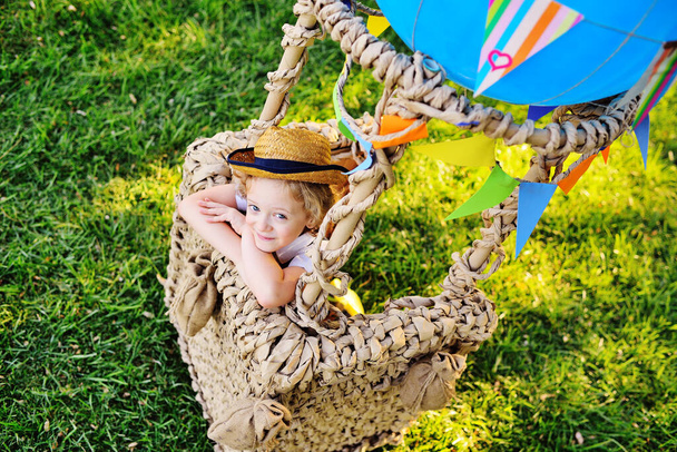 bambino con i capelli ricci in un cappello di paglia seduto in un cesto di un palloncino blu sorride sullo sfondo dell'erba - Foto, immagini