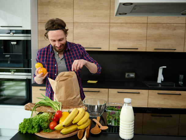 Handsome happy Caucasian man unpacking purchased healthy food standing by a kitchen countertop with bottle of milk and healthy raw vegan vegetables and fruits on a wooden cutting board - Photo, Image