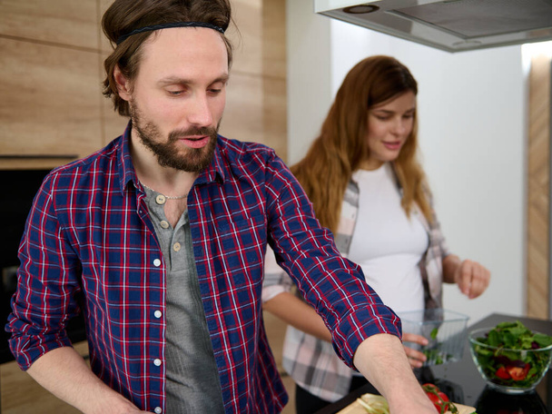 Knappe jonge blanke man in casual kleding bereiden maaltijd met haar zwangere vrouw uit focus op de achtergrond thuis keuken - Foto, afbeelding