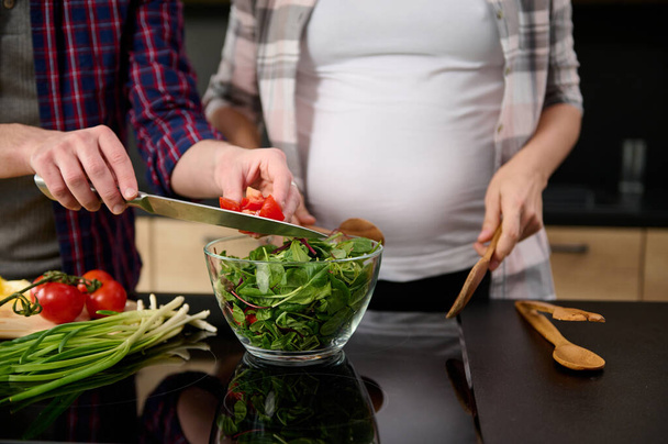 Focus on male hands holding a kitchen knife and puting sliced tomatoes into a bowl with lettuce leaves and greens on the background of his pregnant wife standing by a kitchen island. - Fotó, kép