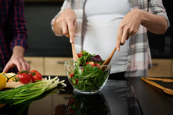 Focus on pregnant woman's hands holding wooden spoons and mixing ingredients in a glass bowl, preparing delicious healthy salad for dinner in the kitchen island next to her husband chopping vegetables - Photo, Image