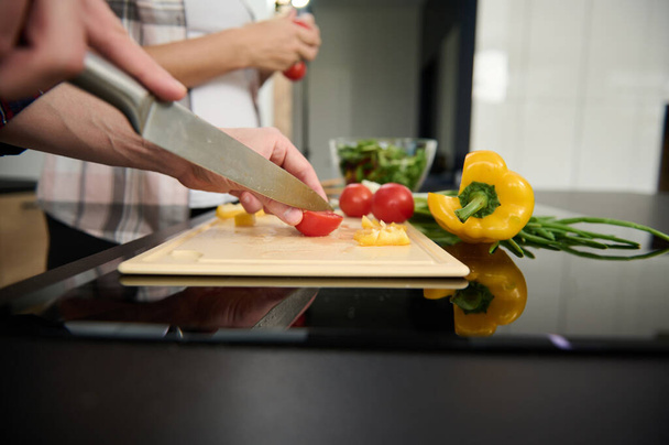 Close-up de mãos de homem cortando tomates e pimentão amarelo em uma tábua de corte no fundo de uma mulher grávida segurando raminho com tomates frescos na cozinha da casa. Conceito de alimentação saudável - Foto, Imagem