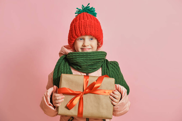 Happy cute caucasian little smiling toddler girl with christmas gift box with red ribbon. girl in a knitted scarf and hat on a pink background,, christmas mood concept copy space - Fotoğraf, Görsel