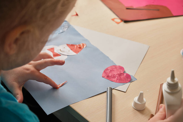 a preschooler girl in a blue T-shirt makes an applique while sitting at the table, a paper snowman hauled on colored paper, children's creativity in winter, atopic dermatitis on the hands of a child - Zdjęcie, obraz